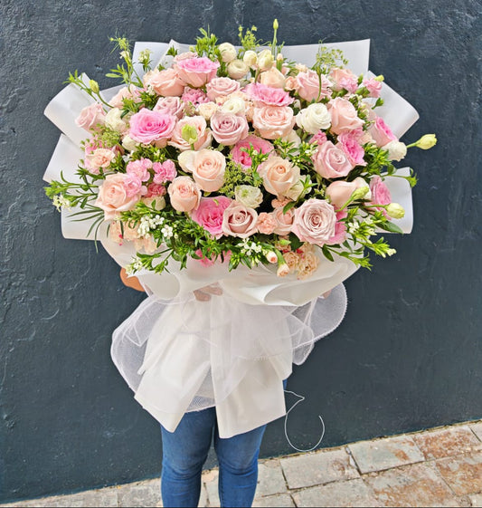 A large, vibrant bouquet of pink and white roses with green foliage, wrapped in white tissue paper and a sheer white bow